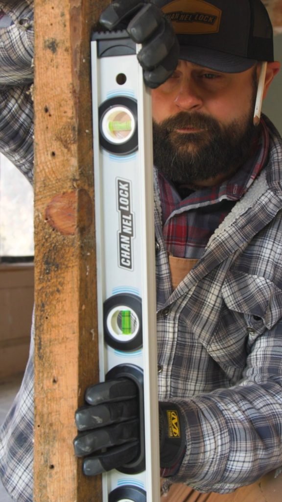 A man in a plaid shirt and gloves holds a CHANNELLOCK® spirit level against a wooden post.