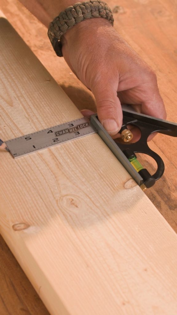 A hand holds a small black tool beside a metal ruler laid along a wooden board.