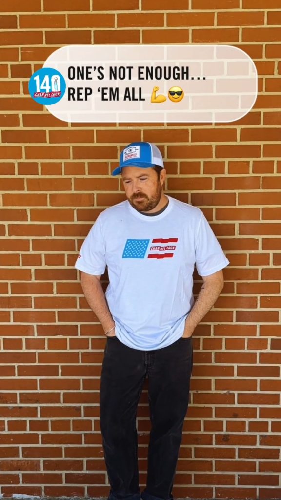 Man wearing a white T-shirt with a small flag graphic and a blue cap stands against a brick wall, with a speech bubble above him reading "ONE'S NOT ENOUGH... REP 'EM ALL'."