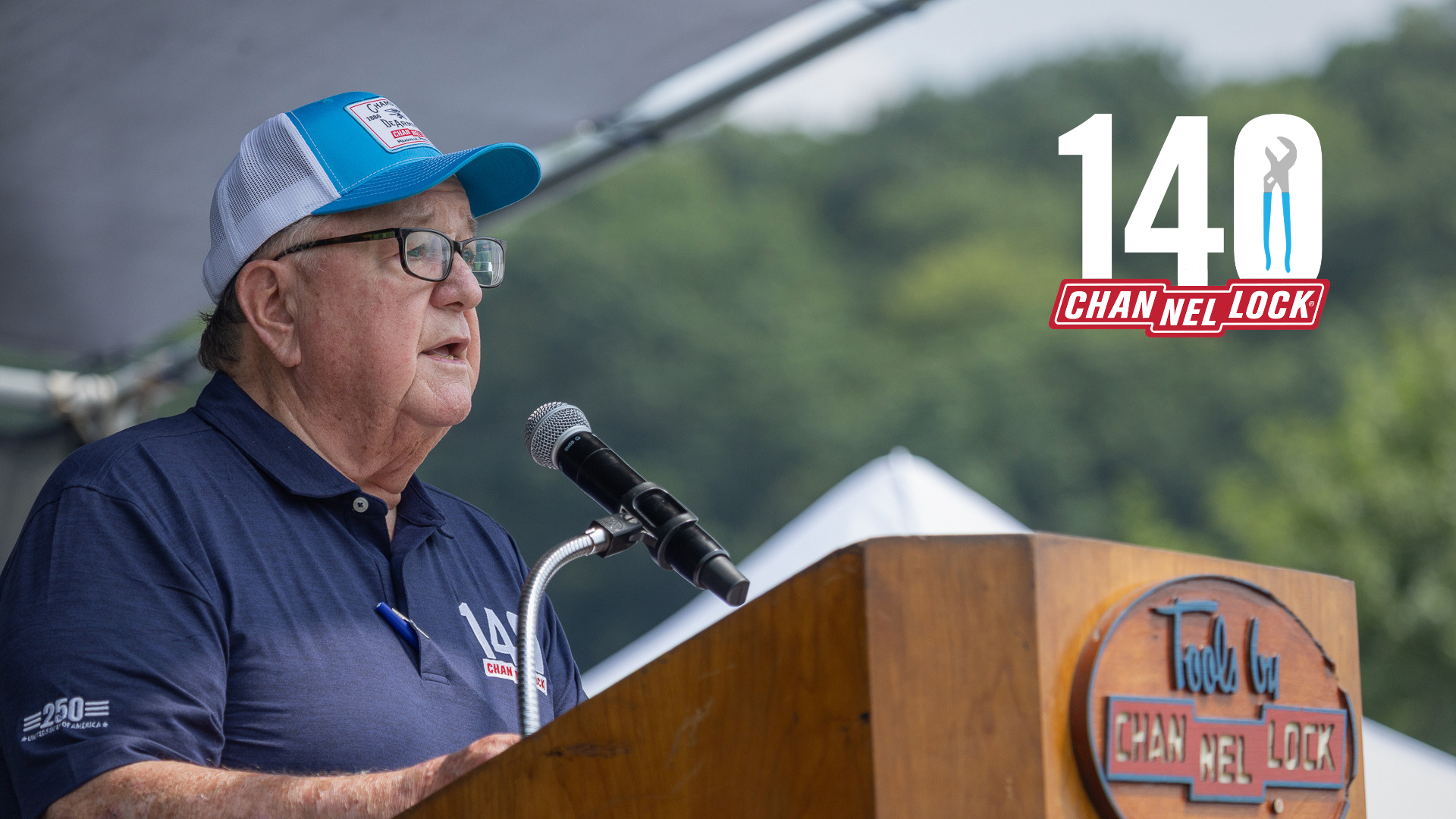 A man wearing a blue polo shirt and blue cap speaks at a CHANNELLOCK® podium with a microphone.