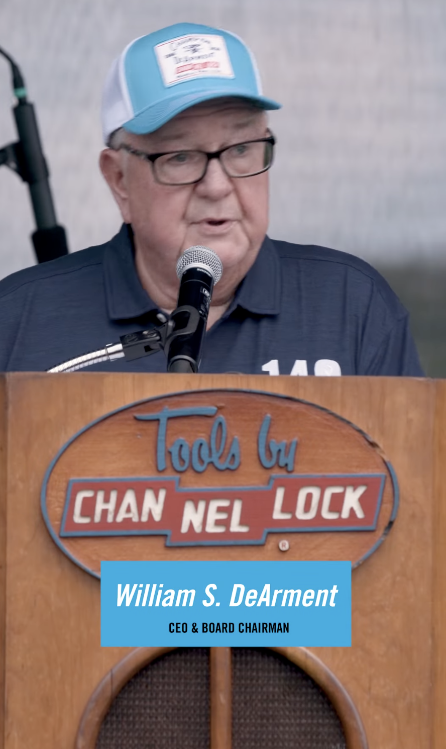 An older man with glasses and a blue cap speaks into a microphone at a CHANNELLOCK® branded podium, with a blue nameplate reading William S. DeArment, CEO & BOARD CHAIRMAN.